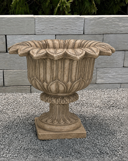 An ornate stone-look planter pot with a fluted pedestal base and wide, scalloped rim, shown outdoors on gravel in front of a light gray stone wall.