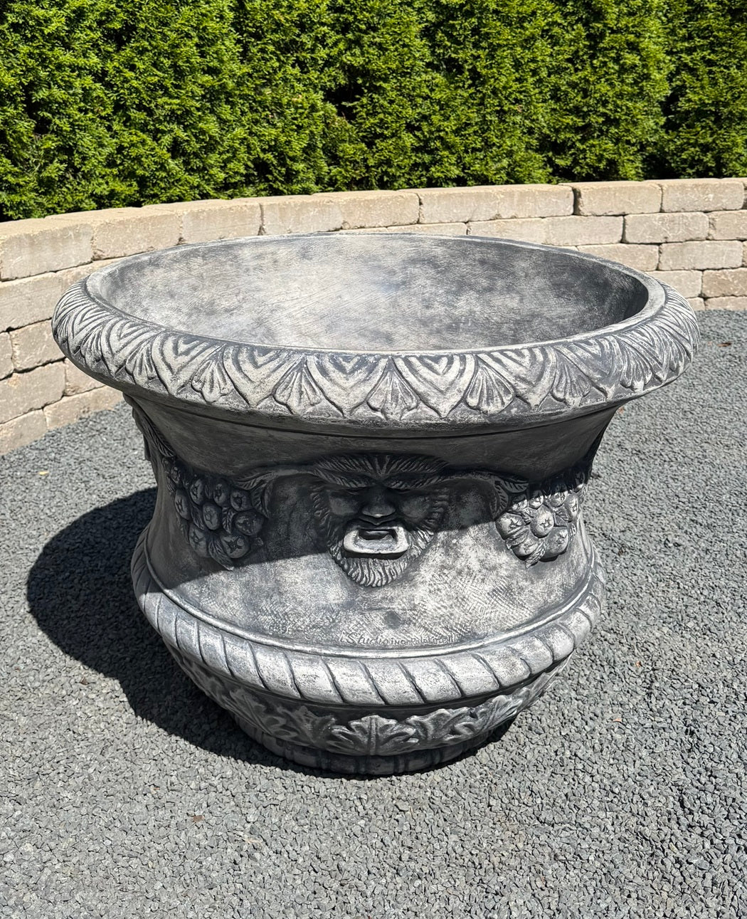 A large gray stone-look decorative planter with carved floral patterns and a sculpted face detail on the front, shown outdoors on gravel with a hedge and low wall in the background.