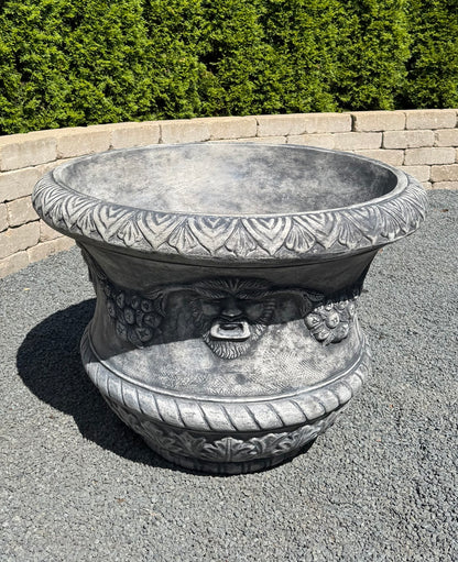 A large gray stone-look decorative planter with carved floral patterns and a sculpted face detail on the front, shown outdoors on gravel with a hedge and low wall in the background.