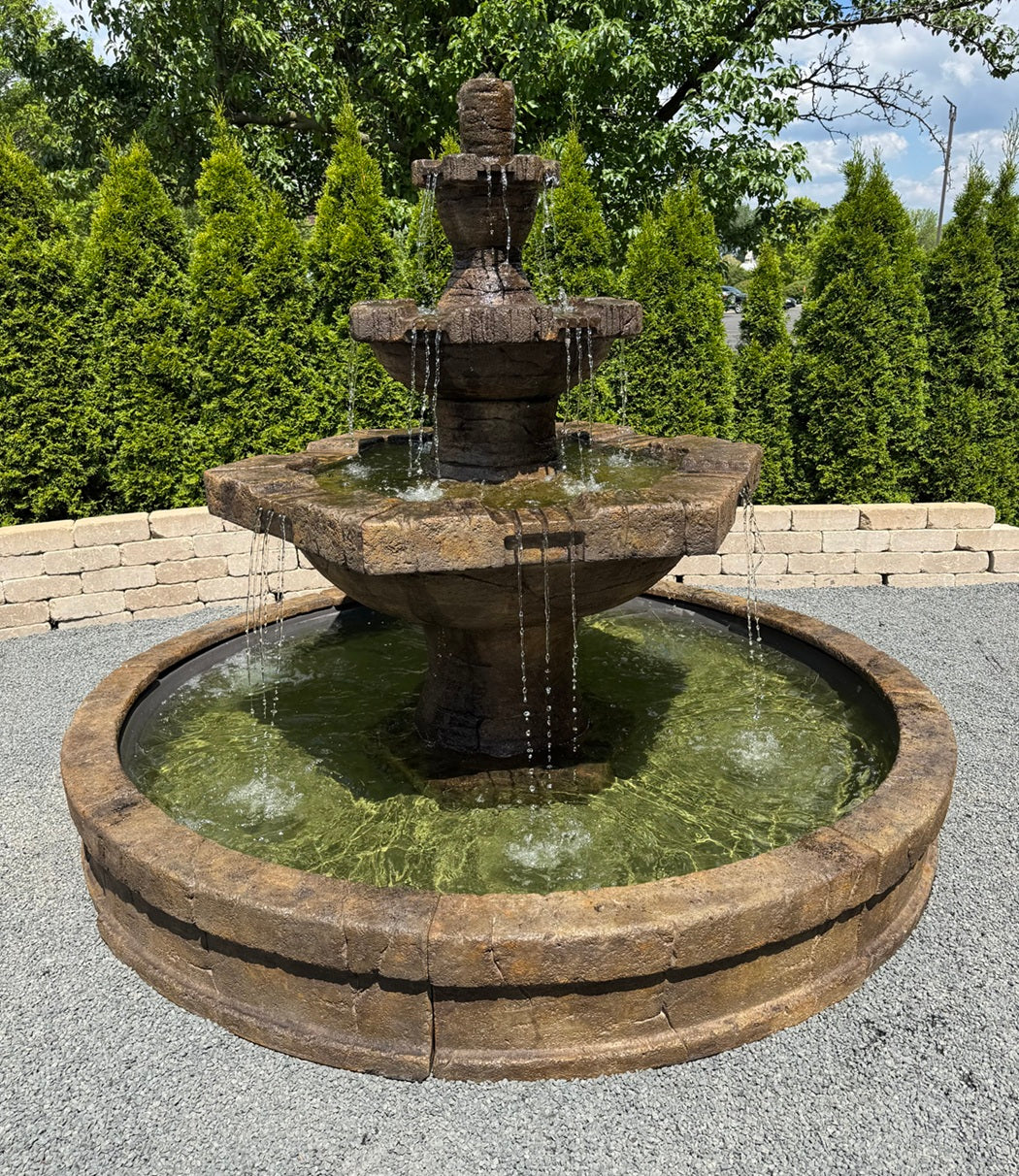 A three-tiered stone fountain with water cascading down, set outdoors on a gravel surface with a stone retaining wall and green trees in the background.