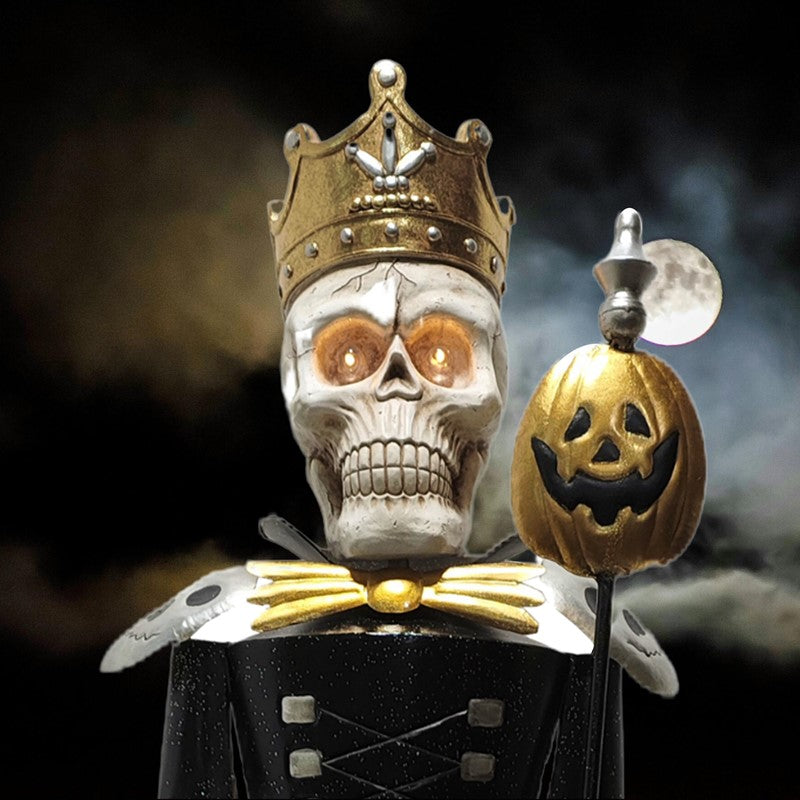 A Halloween decoration shaped like a skull wearing a gold crown and black-and-gold costume, holding a jack-o'-lantern staff against a dark smoky background with a moon visible.