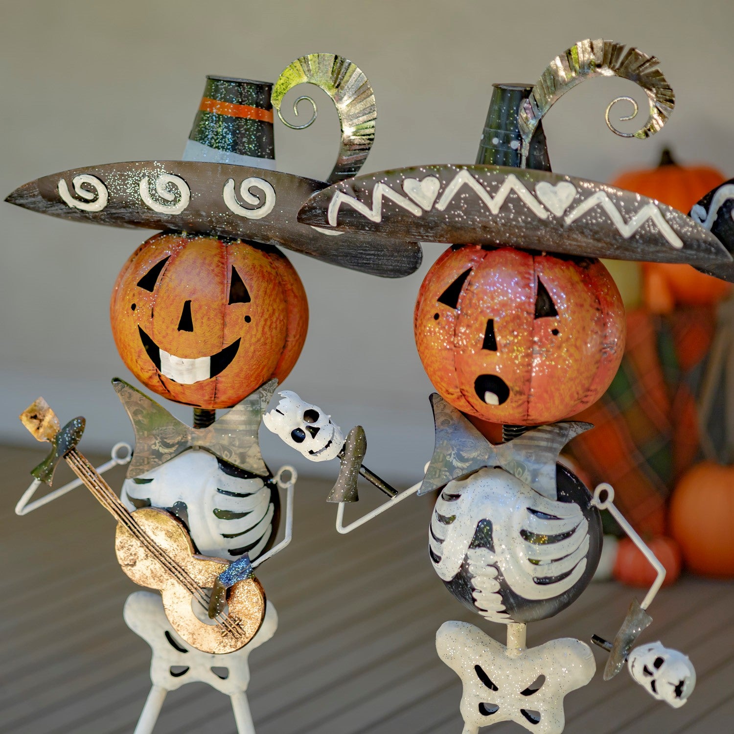 Two Halloween metal skeleton decorations with jack-o'-lantern heads, glittery witch hats, and one holding a guitar and the other holding a small skull, displayed on a tabletop with pumpkins in the background.