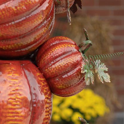Three glossy decorative pumpkins in red, orange, and brown tones with textured ribbing, shown close up outdoors against a blurred brick wall and yellow flowers.