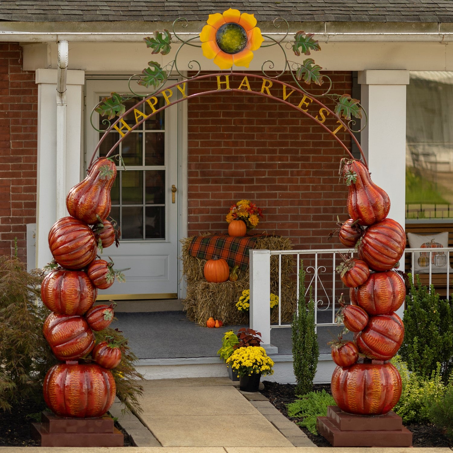 An autumn-themed decorative archway with stacked faux pumpkins on both sides and a sunflower at the top, set in front of a brick house porch with a 'Happy Harvest' sign.