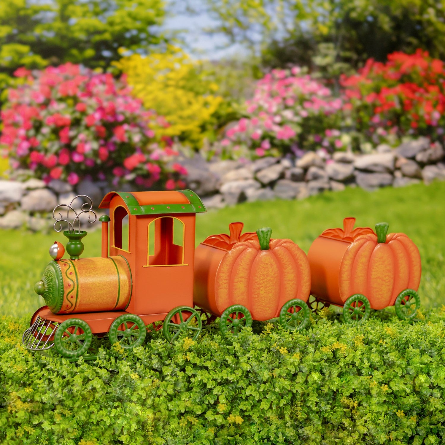 An autumn-themed garden train decoration made from orange and green materials, featuring a small locomotive pulling two pumpkin-shaped cars, displayed on a green hedge with a blurred garden background.
