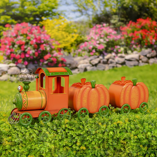 An autumn-themed garden train decoration made from orange and green materials, featuring a small locomotive pulling two pumpkin-shaped cars, displayed on a green hedge with a blurred garden background.