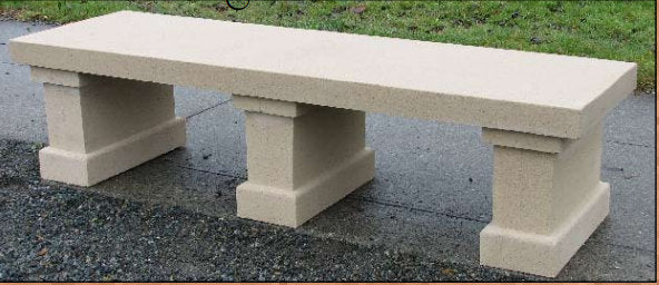 A rectangular concrete bench with block-style legs, shown outdoors on a paved surface with grass in the background.