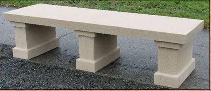 A rectangular concrete bench with block-style legs, shown outdoors on a paved surface with grass in the background.