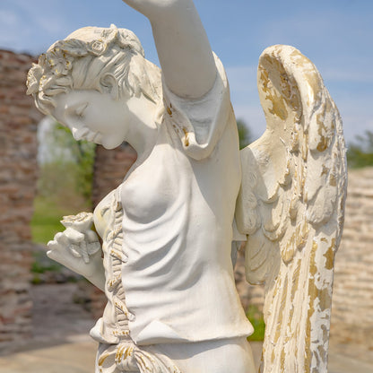 A white angel statue with detailed wings and a classical draped torso, photographed outdoors against a blurred background of stone ruins and blue sky.