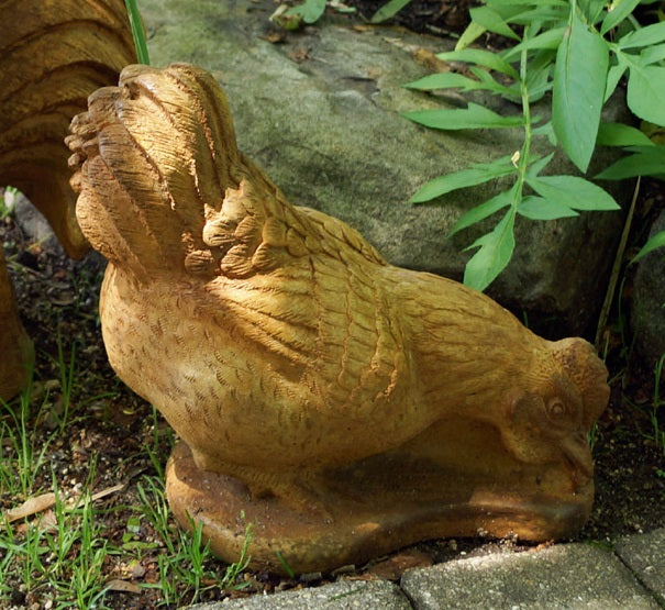 A golden-brown garden statue of a hen pecking at the ground, placed outdoors on a stone patio with green plants and soil in the background.
