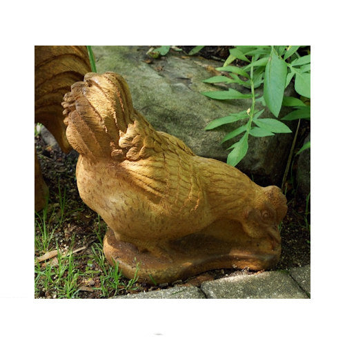 A carved garden statue of a hen pecking rooster in a natural outdoor setting with soil, grass, and green foliage.