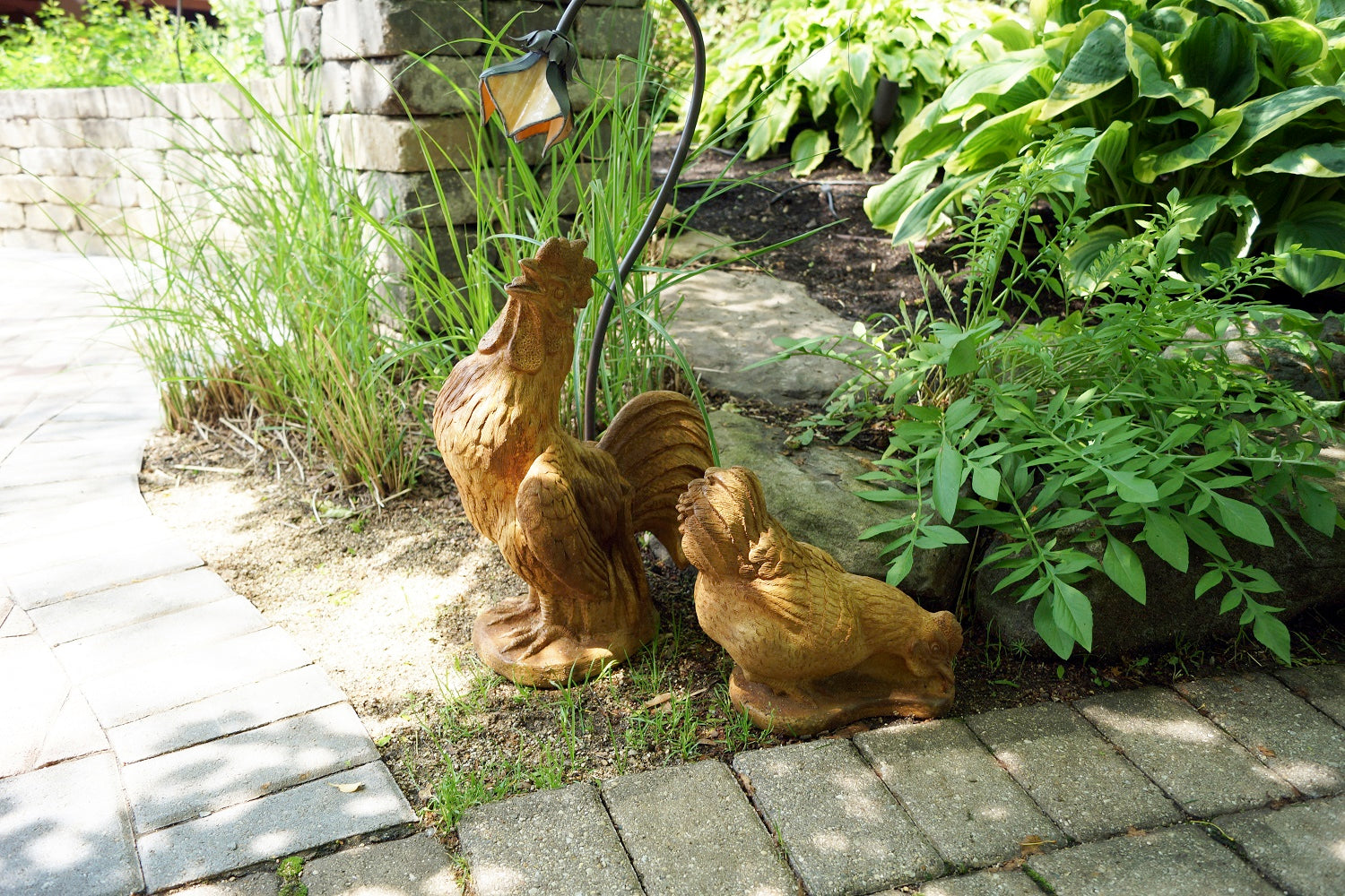 Two rustic brown metal garden statues shaped like a rooster and hen placed on a stone patio beside green plants and grass.