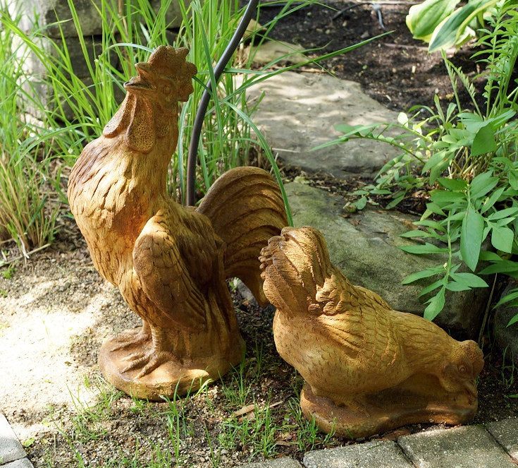 Two decorative garden statues of a rooster and hen in a natural brown wood or stone finish, placed outdoors on a stone path surrounded by green plants.