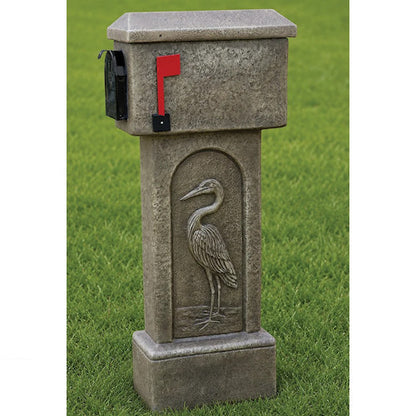 A stone-look mailbox mounted on a matching pedestal post featuring a carved heron design, shown outdoors on a green lawn. The mailbox has a red flag and a black side box.