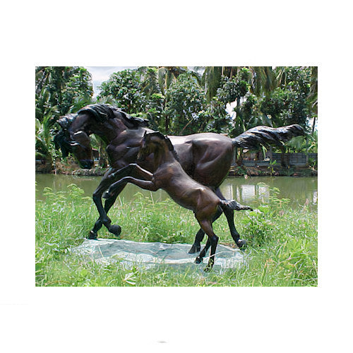 Bronze sculpture of two horses in a grassy area with a body of water and trees in the background.