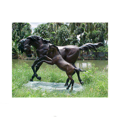 Bronze sculpture of two horses in a grassy area with a body of water and trees in the background.