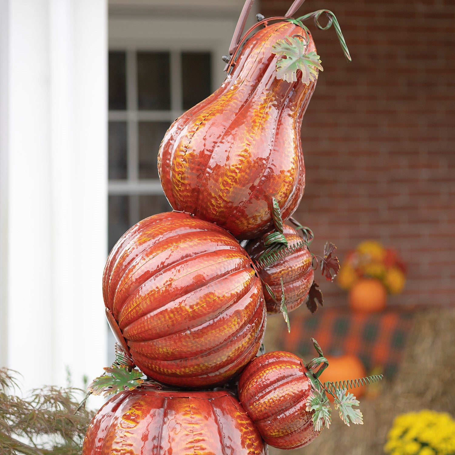 A decorative stack of faux gourds and pumpkins in orange, red, and brown tones with glossy textured finishes and small green leaf accents, photographed outdoors against a blurred brick wall and window background.