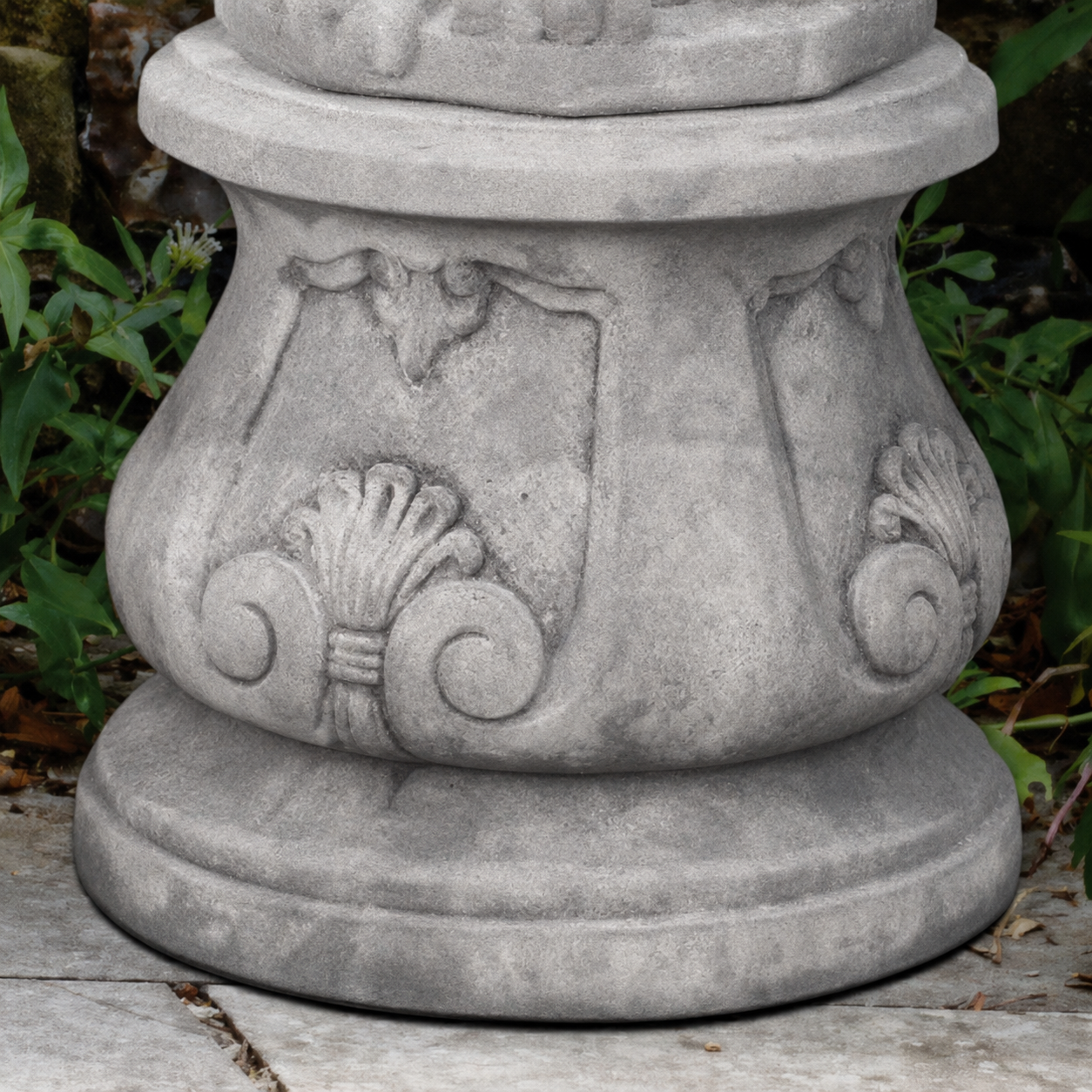 Gray stone garden pedestal with carved decorative scrollwork and shell motifs, shown outdoors on a stone surface with green foliage in the background.