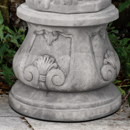 Gray stone garden pedestal with carved decorative scrollwork and shell motifs, shown outdoors on a stone surface with green foliage in the background.
