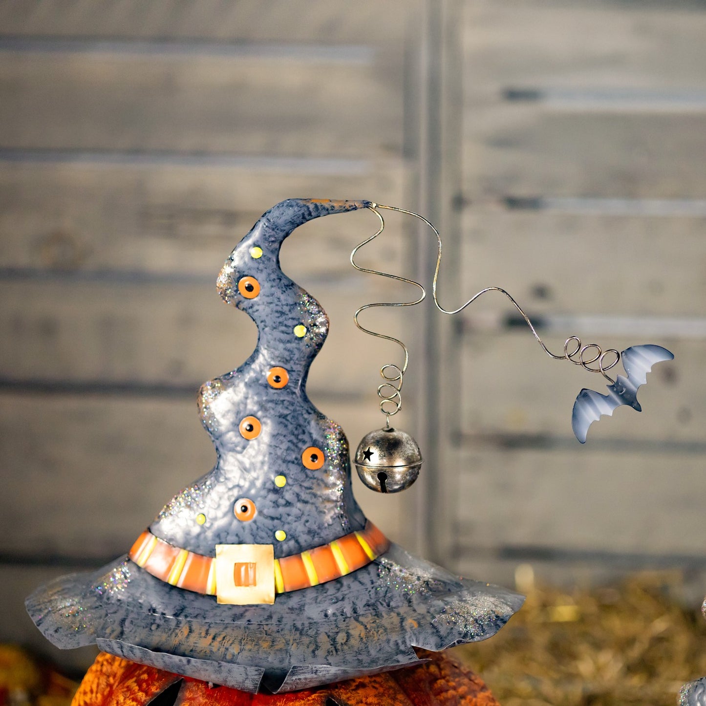 A decorative metal witch hat ornament with a silver bell and wire tail, displayed on a pumpkin against a rustic wooden background.