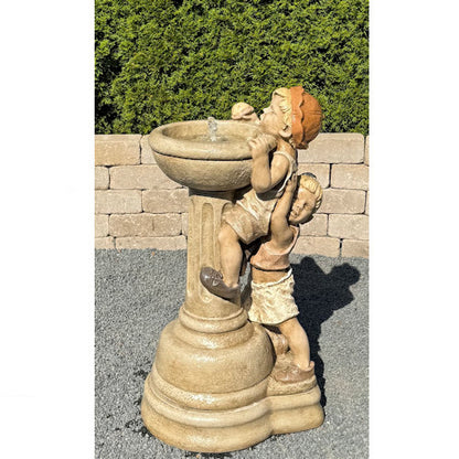 A beige stone-style garden fountain sculpture featuring two children climbing and drinking from a pedestal basin, photographed outdoors against a hedge and brick wall.
