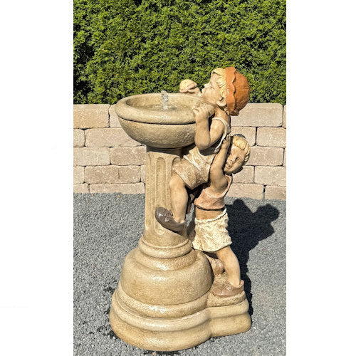 A beige stone-style garden fountain sculpture featuring two children climbing and drinking from a pedestal basin, photographed outdoors against a hedge and brick wall.