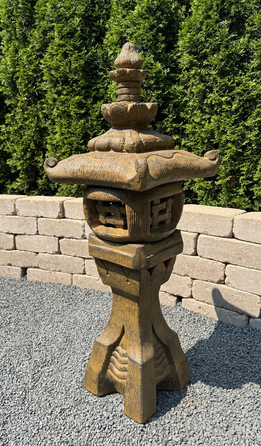 A carved stone Japanese-style garden lantern standing on a gravel surface with a stone wall and green hedge in the background.