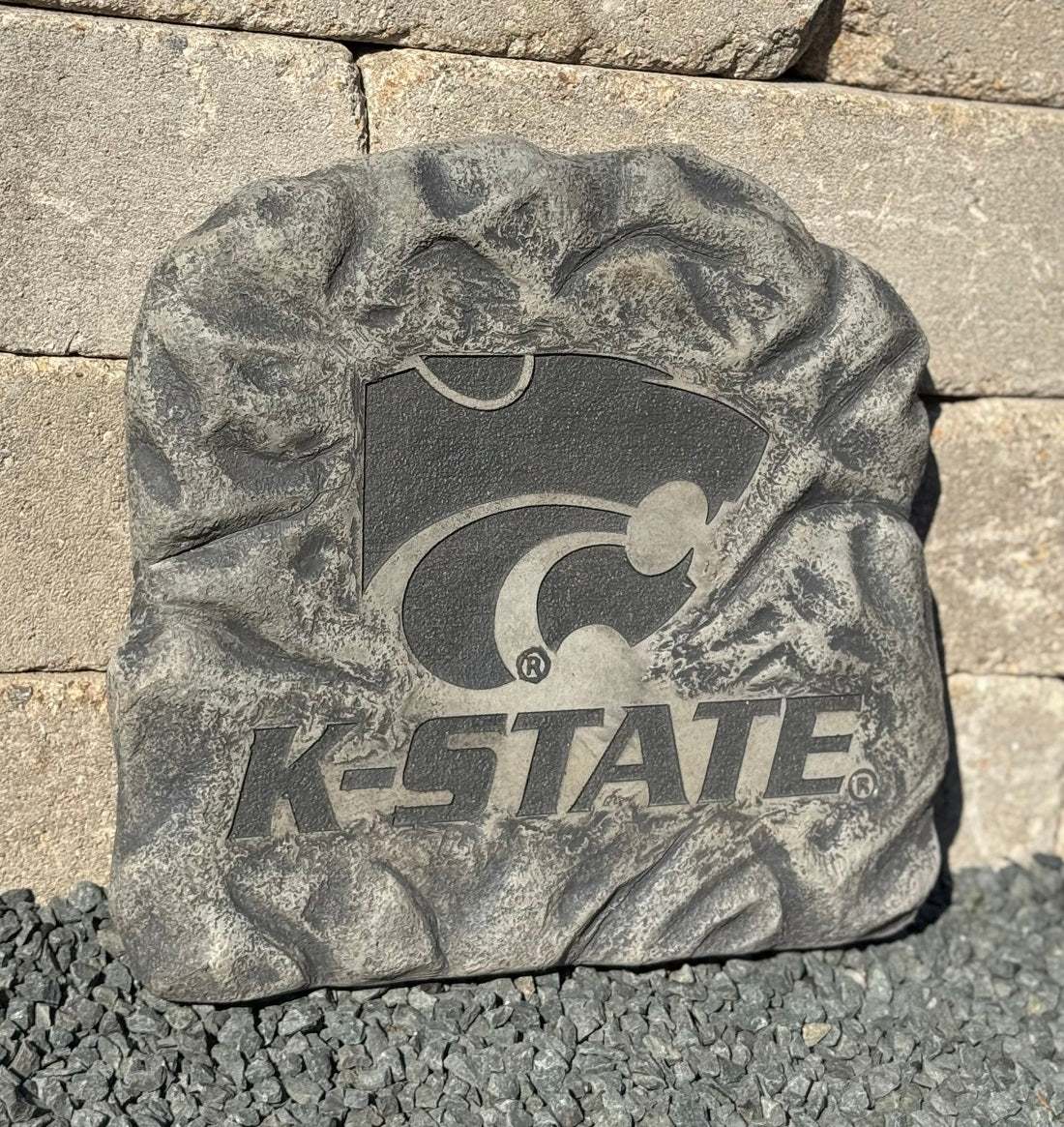 A textured gray decorative stone plaque featuring the K-State Wildcats logo and 'K-STATE' text, photographed outdoors against a stone wall and gravel surface.