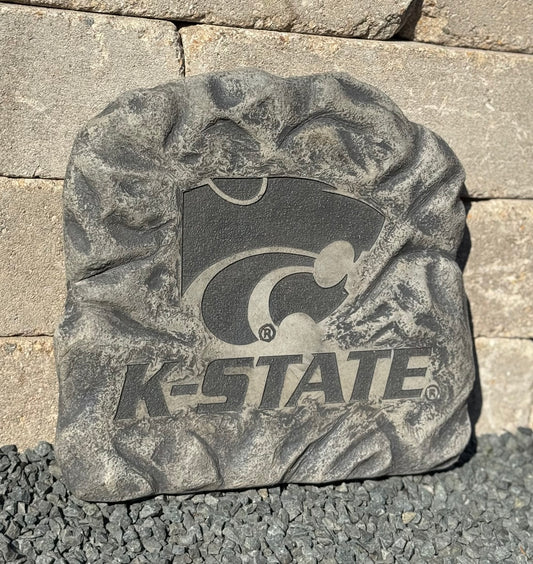 A textured gray decorative stone plaque featuring the K-State Wildcats logo and 'K-STATE' text, photographed outdoors against a stone wall and gravel surface.