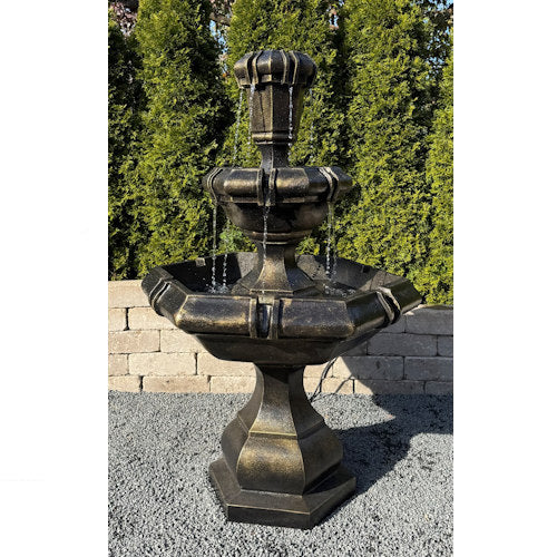A three-tiered outdoor stone fountain with a distressed bronze finish, shown on a gravel surface with a brick wall and green hedges in the background.
