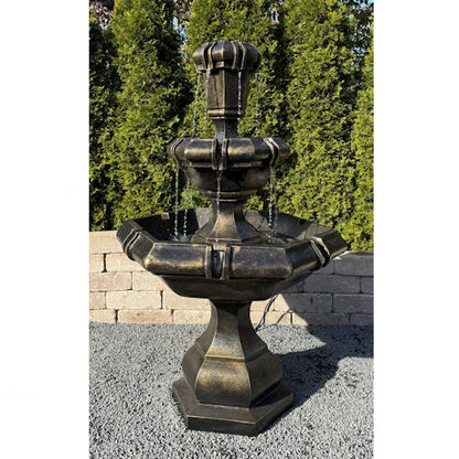 A three-tiered outdoor stone fountain with a distressed bronze finish, shown on a gravel surface with a brick wall and green hedges in the background.