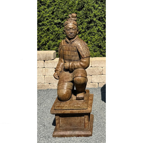 A carved terracotta warrior garden statue in a kneeling pose, mounted on a rectangular pedestal, photographed outdoors against a hedge and stone wall.