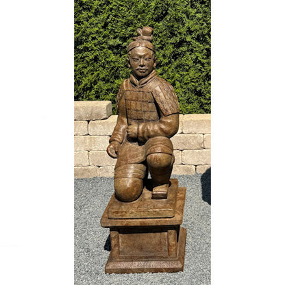 A carved terracotta warrior garden statue in a kneeling pose, mounted on a rectangular pedestal, photographed outdoors against a hedge and stone wall.