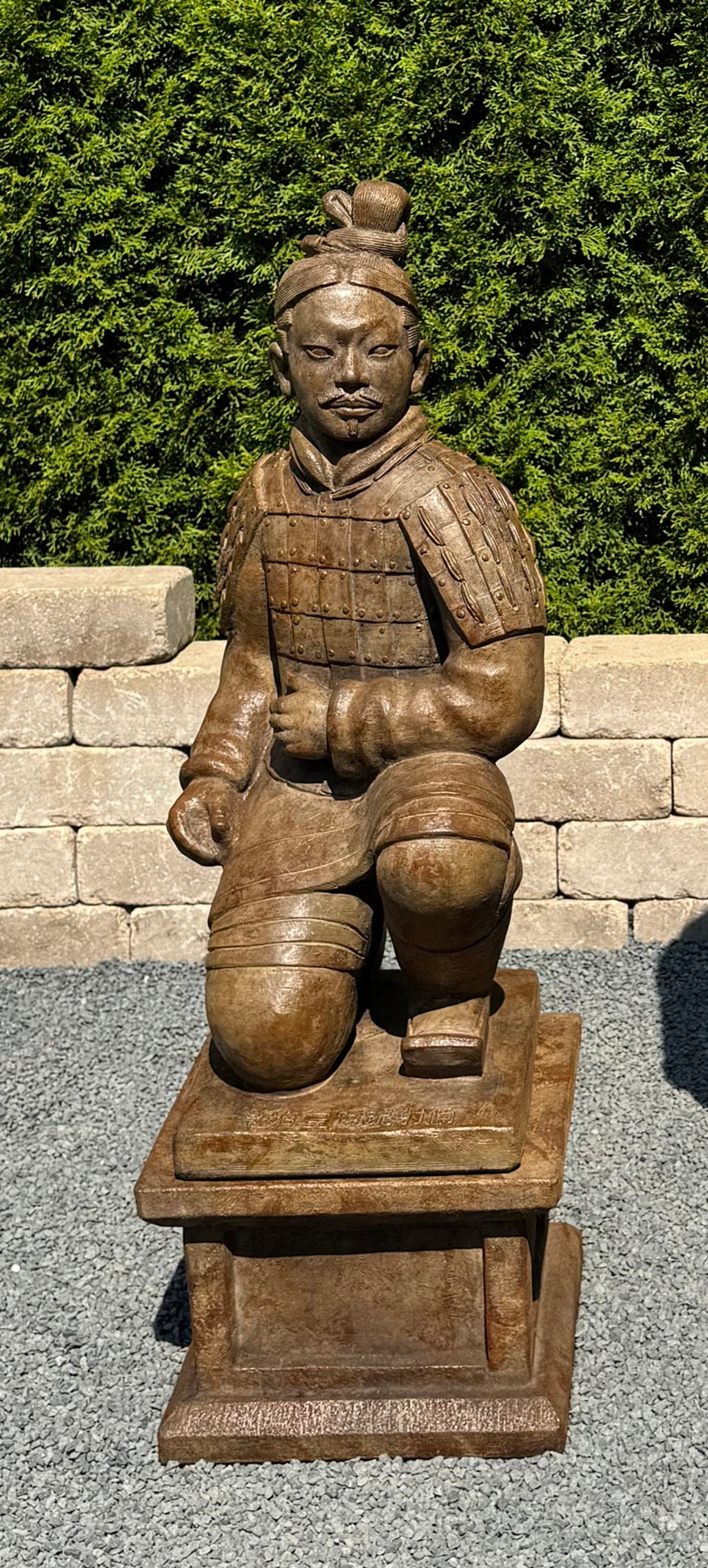 A bronze-colored terracotta warrior statue kneeling on a rectangular pedestal, photographed outdoors against a background of green hedges and a light stone wall.