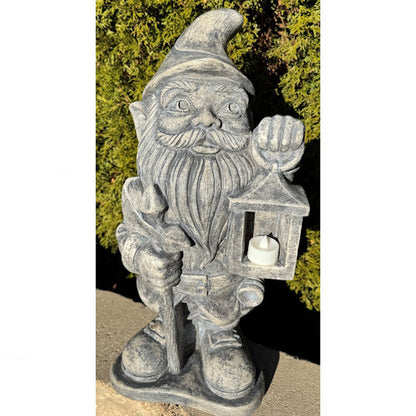 A gray stone garden gnome figurine holding a lantern with a white tea light candle inside, photographed outdoors against green foliage.