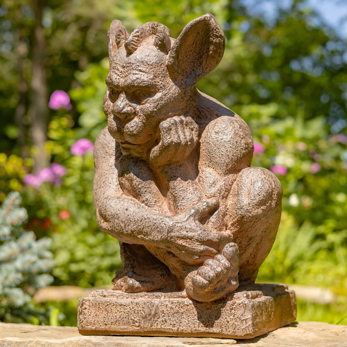 A small stone gargoyle garden statue in a crouching pose, shown outdoors on a stone surface with a blurred green garden background.