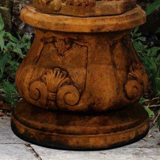 Close-up of a terracotta-colored stone or concrete garden pedestal base with carved scroll and shell-like decorative details, shown outdoors on a stone surface with green plants in the background.