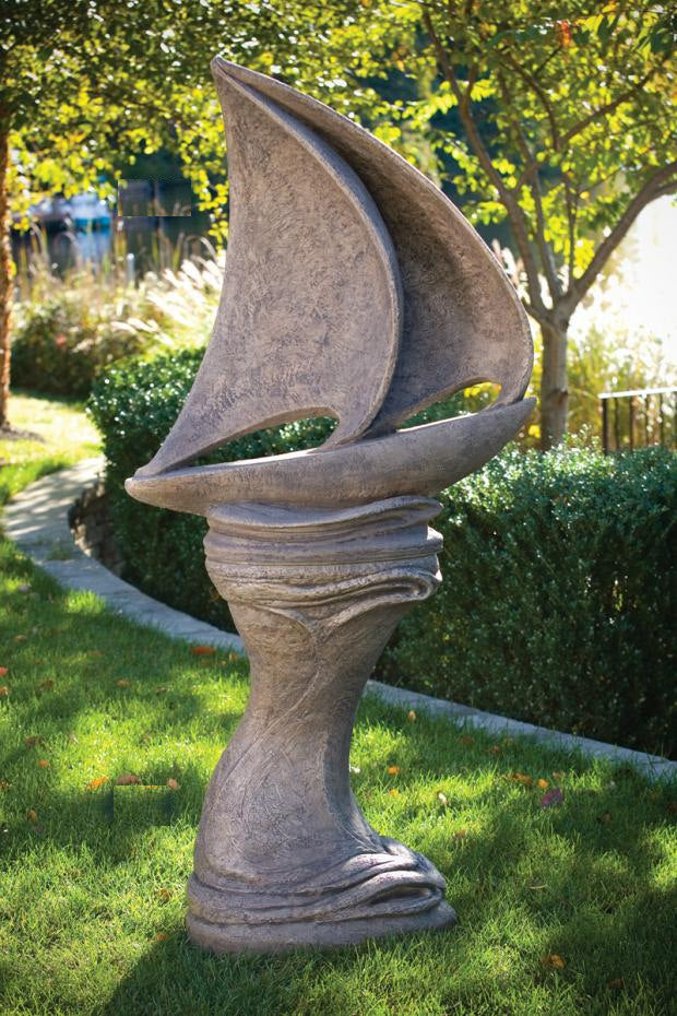A large abstract stone garden sculpture shaped like a stylized sailboat or curved pedestal form, placed outdoors on a grassy lawn with hedges and trees in the background.