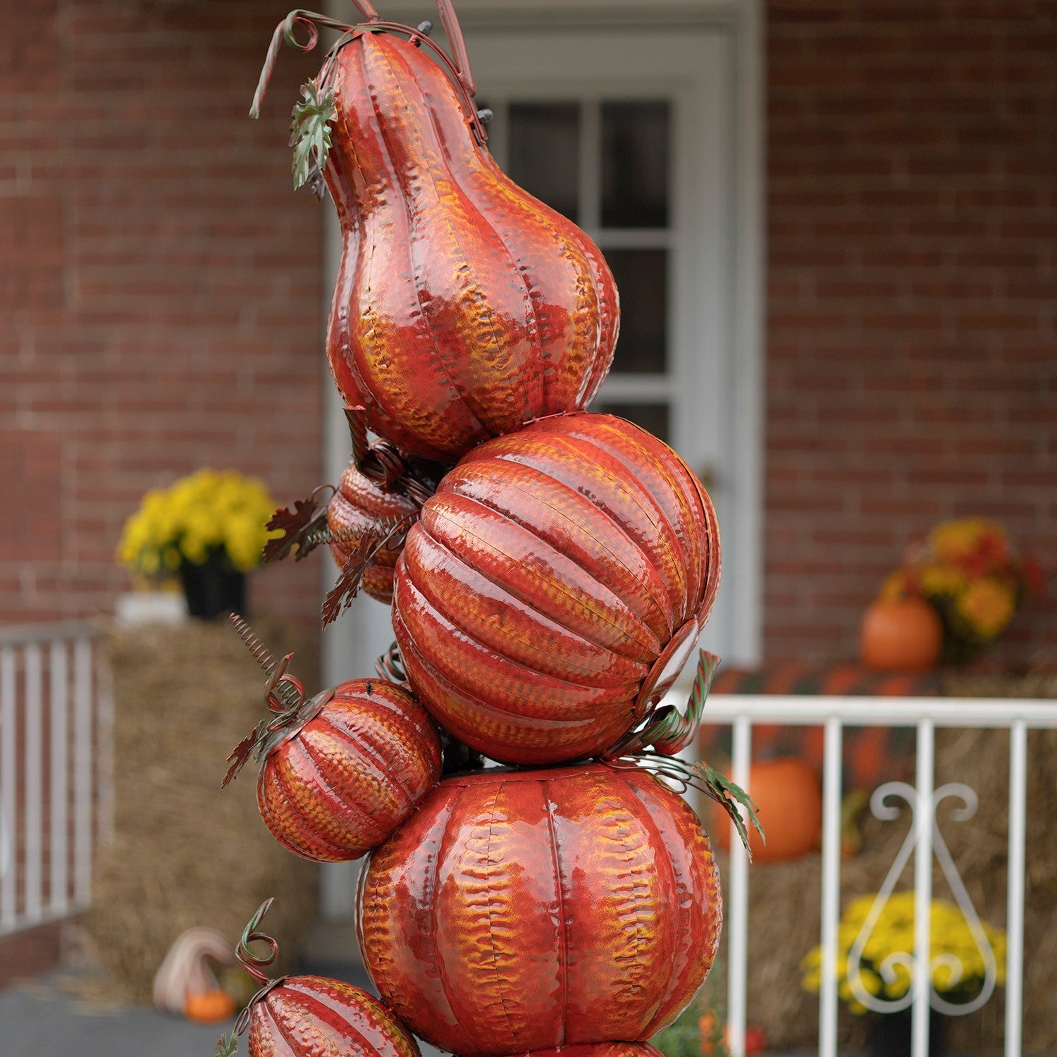 A decorative autumn hanging pumpkin tree decoration made of stacked faux gourds in orange, red, and brown tones, shown hanging in front of a brick house porch.