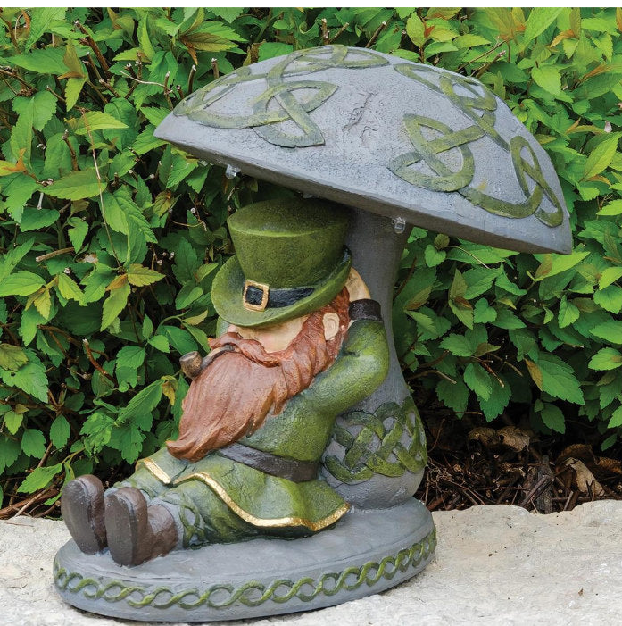 A decorative garden statue of a seated leprechaun under a mushroom-shaped roof, placed outdoors in front of green foliage. The leprechaun wears a green hat and coat with a reddish-brown beard, and the mushroom cap has green Celtic knot detailing.