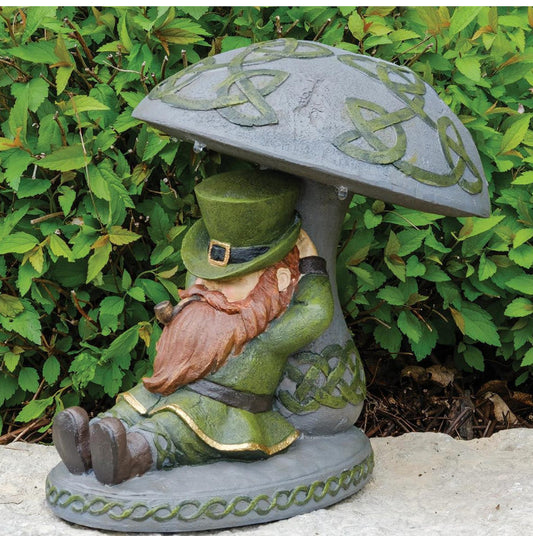 A decorative garden statue of a seated leprechaun under a mushroom-shaped roof, placed outdoors in front of green foliage. The leprechaun wears a green hat and coat with a reddish-brown beard, and the mushroom cap has green Celtic knot detailing.