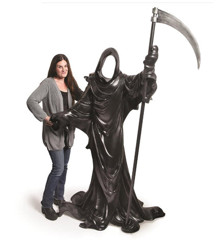 A large black grim reaper statue holding a scythe stands next to a woman with long dark hair wearing a gray cardigan, black top, and blue jeans, against a plain white background.