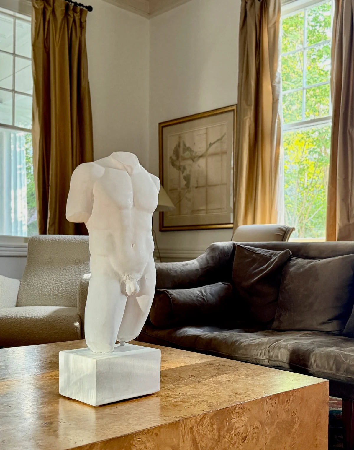 A white classical-style male torso sculpture on a square pedestal base, displayed on a wooden table in a living room with a sofa, curtains, and windows in the background