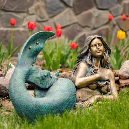 Bronze mermaid statue with a tail in a garden setting with tulips and rocks.