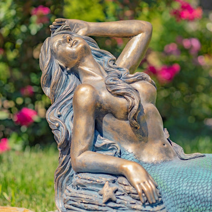 Bronze mermaid statue with green grass and pink flowers in the background