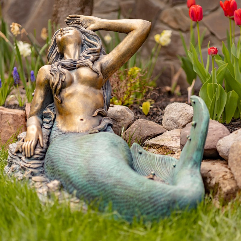 Bronze mermaid statue in a garden setting with flowers and rocks.