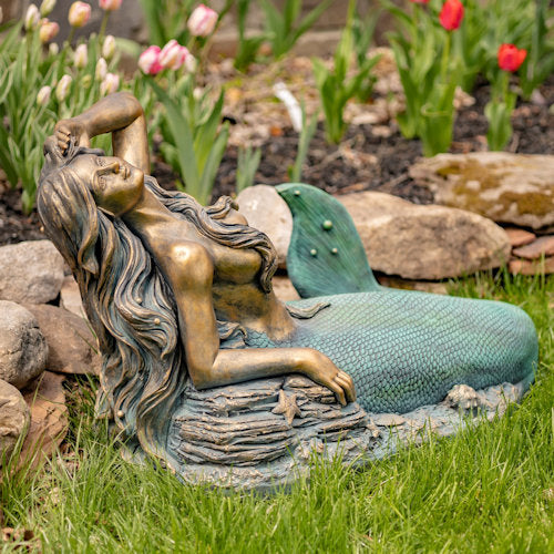 Bronze mermaid statue in a garden setting with flowers and rocks.
