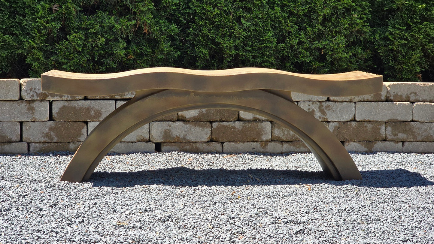 A modern curved concrete bench with a wavy top and arched base, situated outdoors on a gravel surface in front of a stone wall and green hedges.