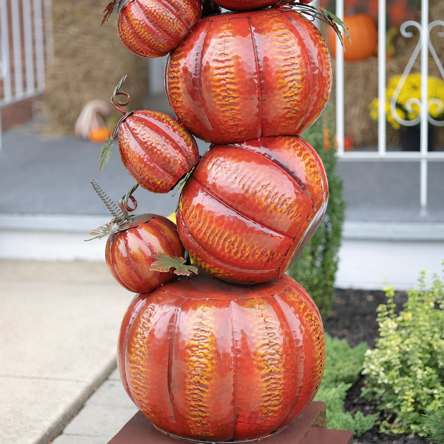 A decorative stack of glossy orange and red faux pumpkins with textured ribbing and small green stems, displayed outdoors on a pedestal in a garden setting.