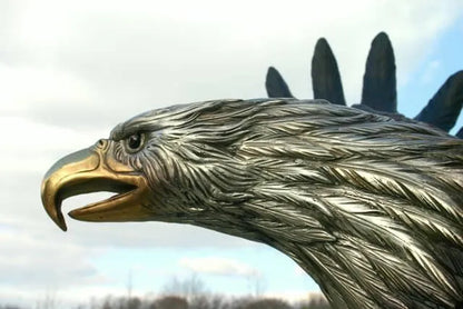 Close-up of a metallic eagle head sculpture or statue, showing detailed feather texture, a gold-toned beak, and dark wingtips against an outdoor sky background.